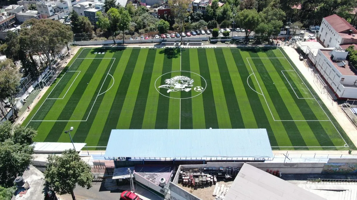 Vista aérea de una cancha de fútbol 7 con pasto sintético y logotipo al centro junto a un edificio deportivo