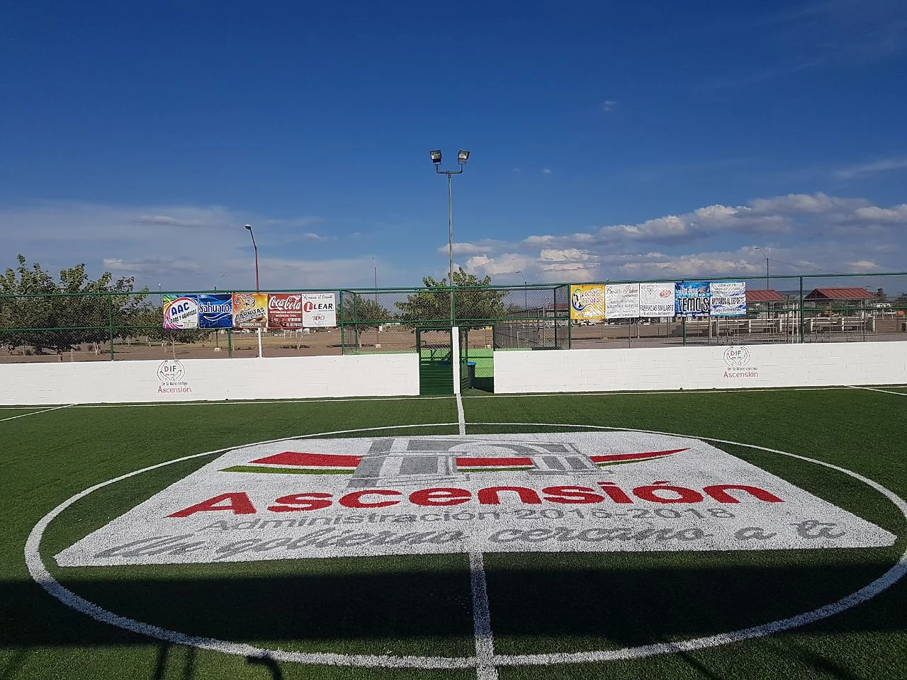 Logotipo del municipio de Ascensión, Chihuahua, insertado en pasto sintético blanco, rojo y verde en el círculo central de una cancha de fútbol rápido