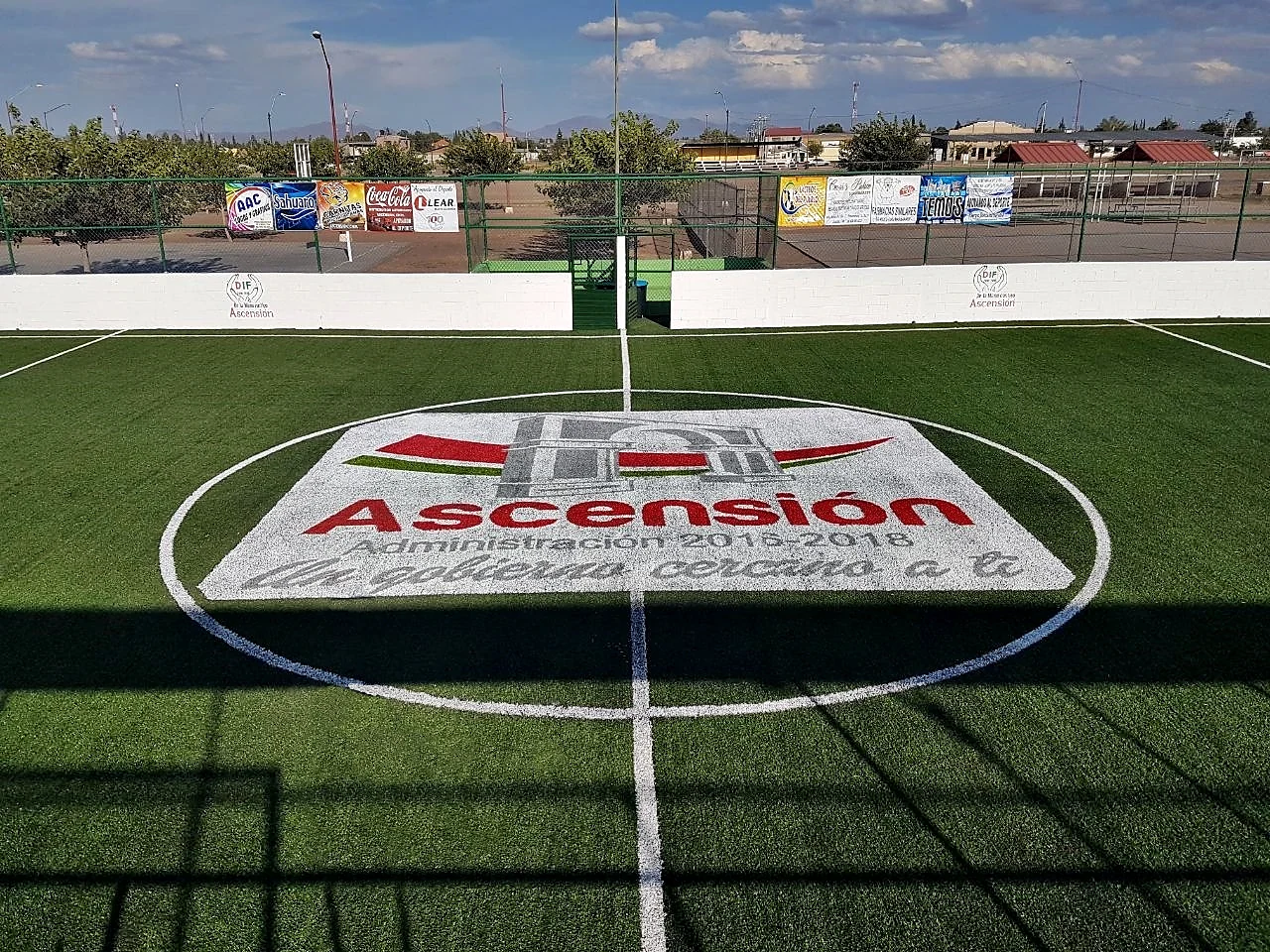 Vista desde la línea media del logotipo municipal de Ascensión insertado en el pasto sintético de una unidad deportiva