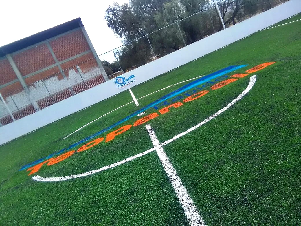 Nombre Teopancala en pasto sintético azul y naranja insertado sobre el círculo central de una cancha, visto en diagonal