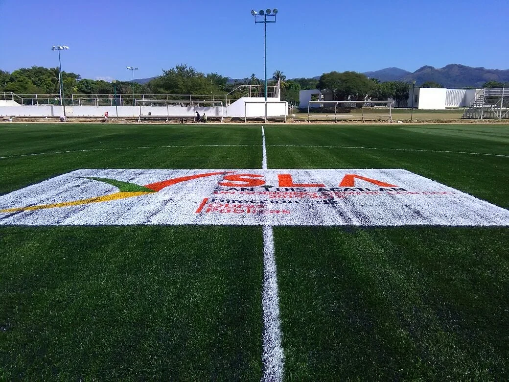 Vista general de una cancha de pasto sintético con el logotipo SLA insertado en el círculo central y postes de alumbrado