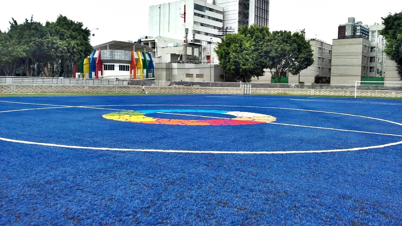 Vista general de una cancha de pasto sintético azul con un logotipo multicolor en el círculo central y edificios al fondo en la Ciudad de México