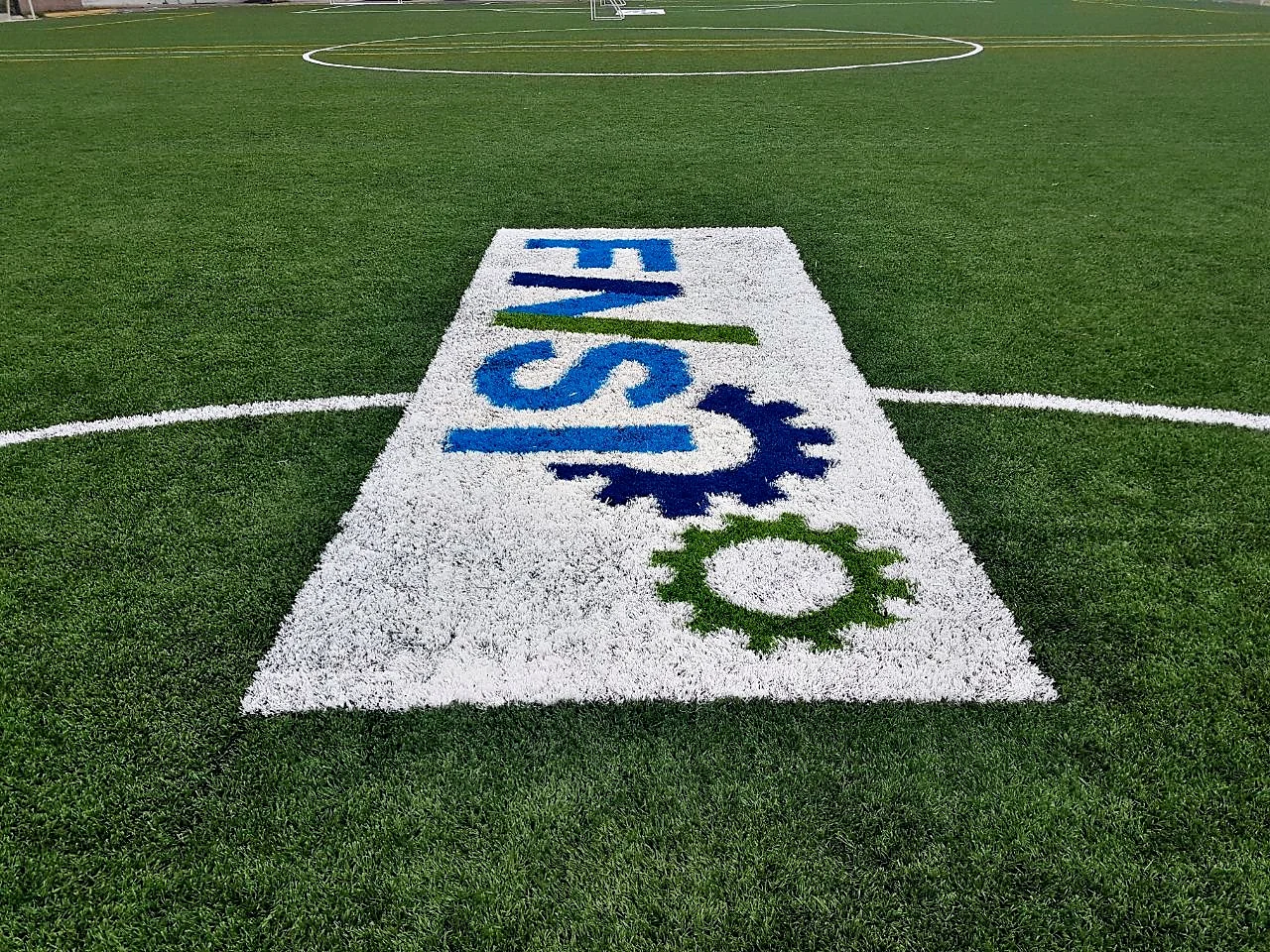Logotipo FNSI con engranes insertado en pasto sintético en el círculo central de una cancha en Apodaca, Nuevo León, vista frontal