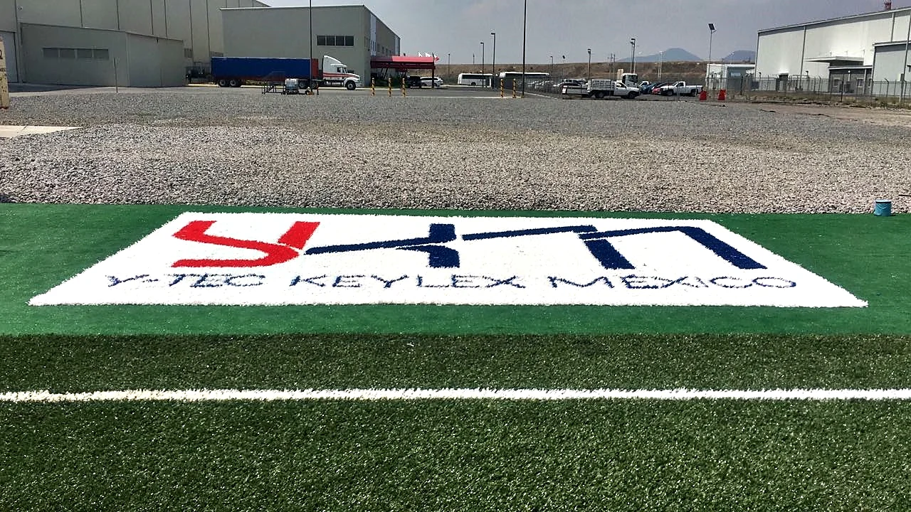Logotipo Y-Tec Keylex México insertado en pasto sintético, vista frontal con la planta al fondo
