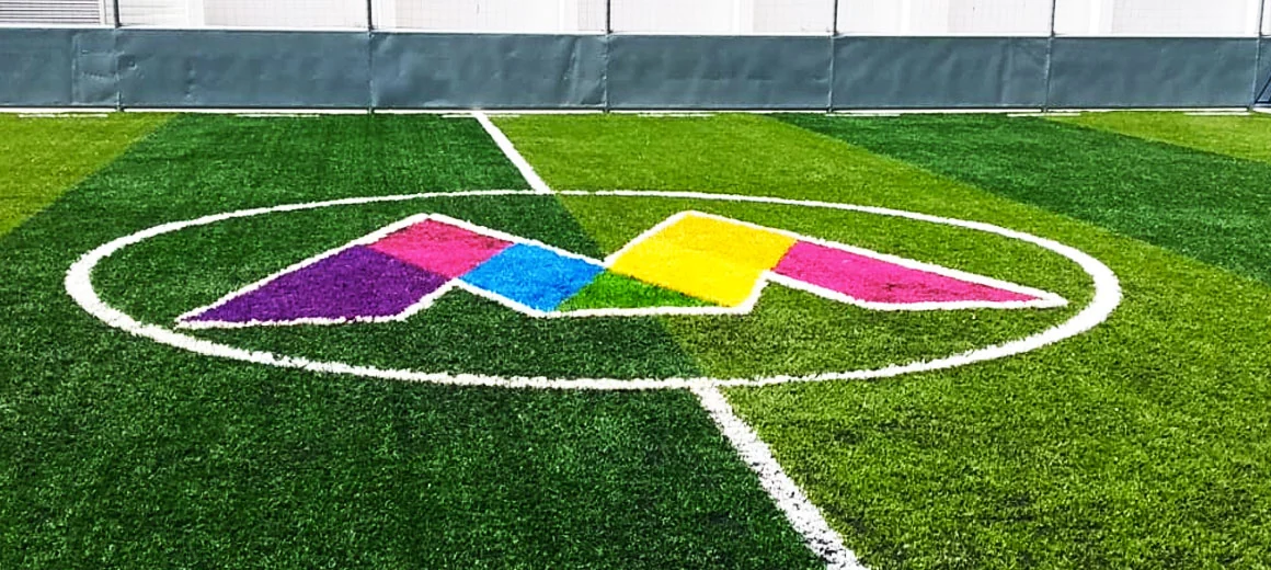 Logotipo multicolor en morado, verde, amarillo y rosa insertado en pasto sintético en el círculo central de una cancha de fútbol rápido