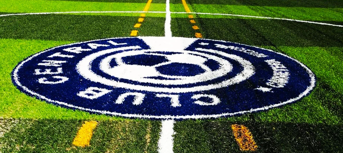 Escudo circular del Club Central en azul y blanco con un balón al centro, insertado en pasto sintético en el círculo central de una cancha con líneas amarillas de un segundo deporte