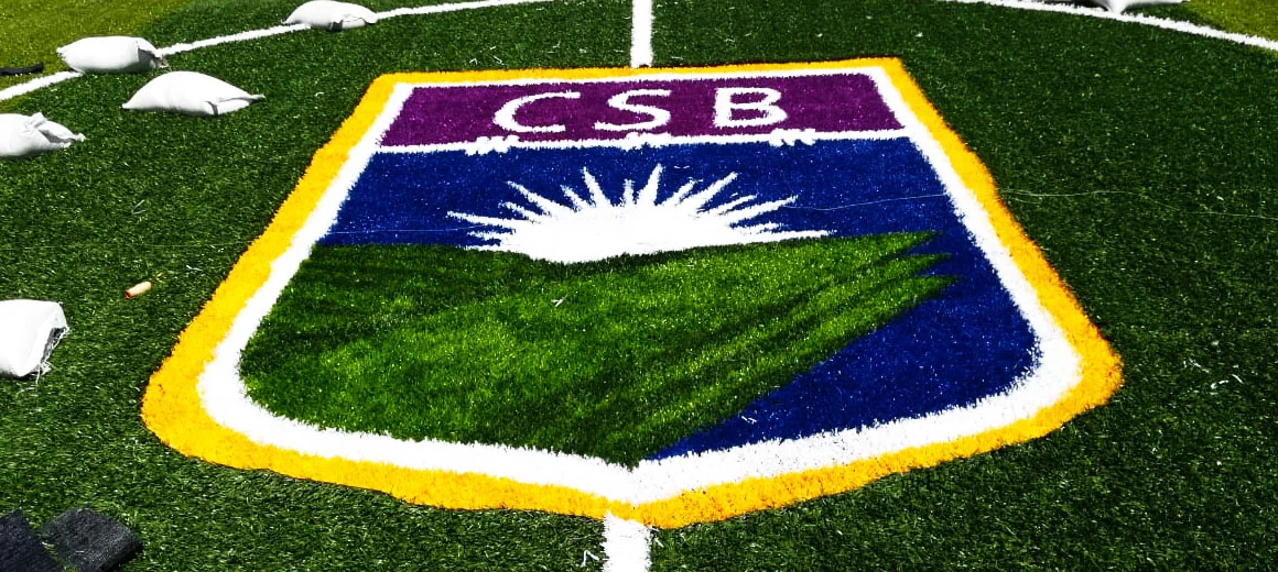 Escudo CSB en morado, amarillo, verde y blanco insertado en pasto sintético junto a la línea lateral de la cancha