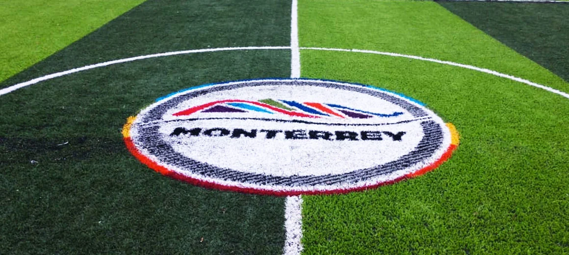 Logotipo de la ciudad de Monterrey con anillo blanco y cerro multicolor insertado en pasto sintético en el círculo central de una cancha de fútbol
