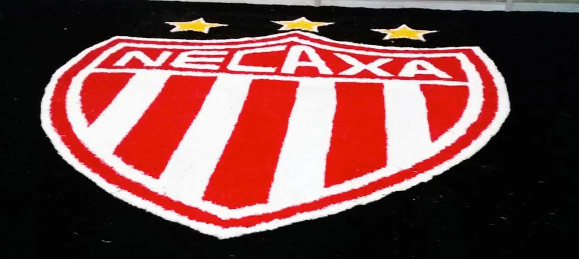 Escudo del Club Necaxa en rojo y blanco con tres estrellas amarillas armado en pasto sintético sobre piso de taller antes de instalarse en la cancha