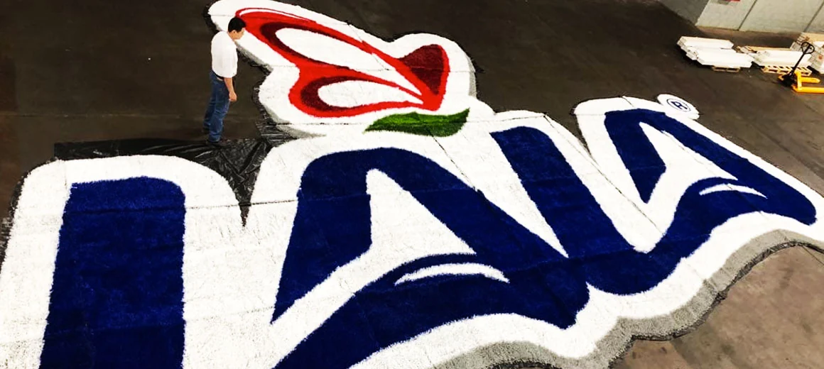 Logotipo LALA en azul, blanco y rojo armado en pasto sintético en taller, con un integrante de la cuadrilla que da la escala del tamaño