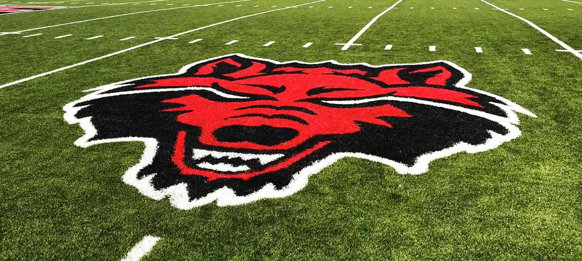 Cabeza de lobo en rojo y negro insertada en pasto sintético entre las líneas de yarda del campo de fútbol americano de Arkansas State University, Querétaro