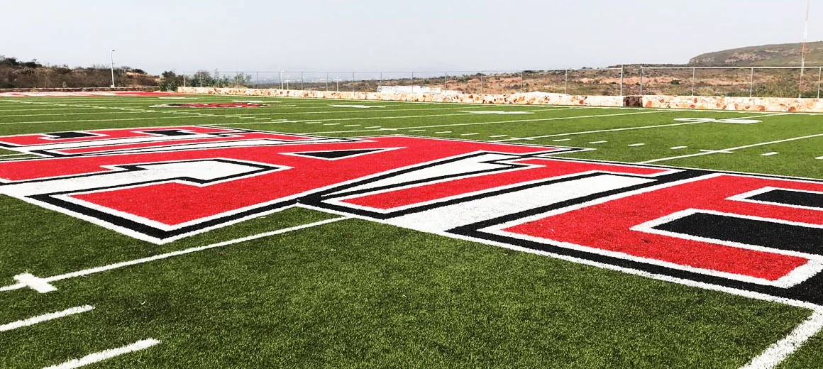 Letras STATE en rojo y negro insertadas en pasto sintético en la zona de anotación del campo de fútbol americano de Arkansas State University, Querétaro, vistas en diagonal