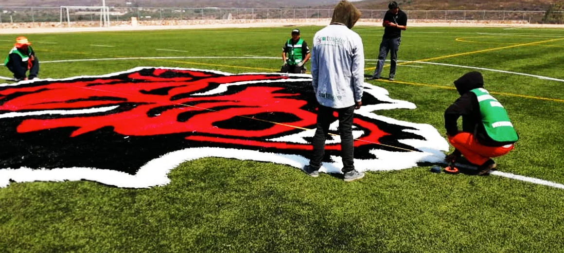 Cabeza de lobo insertada en el campo de fútbol americano de Arkansas State University, Querétaro, con la cuadrilla terminando el relleno alrededor del logotipo