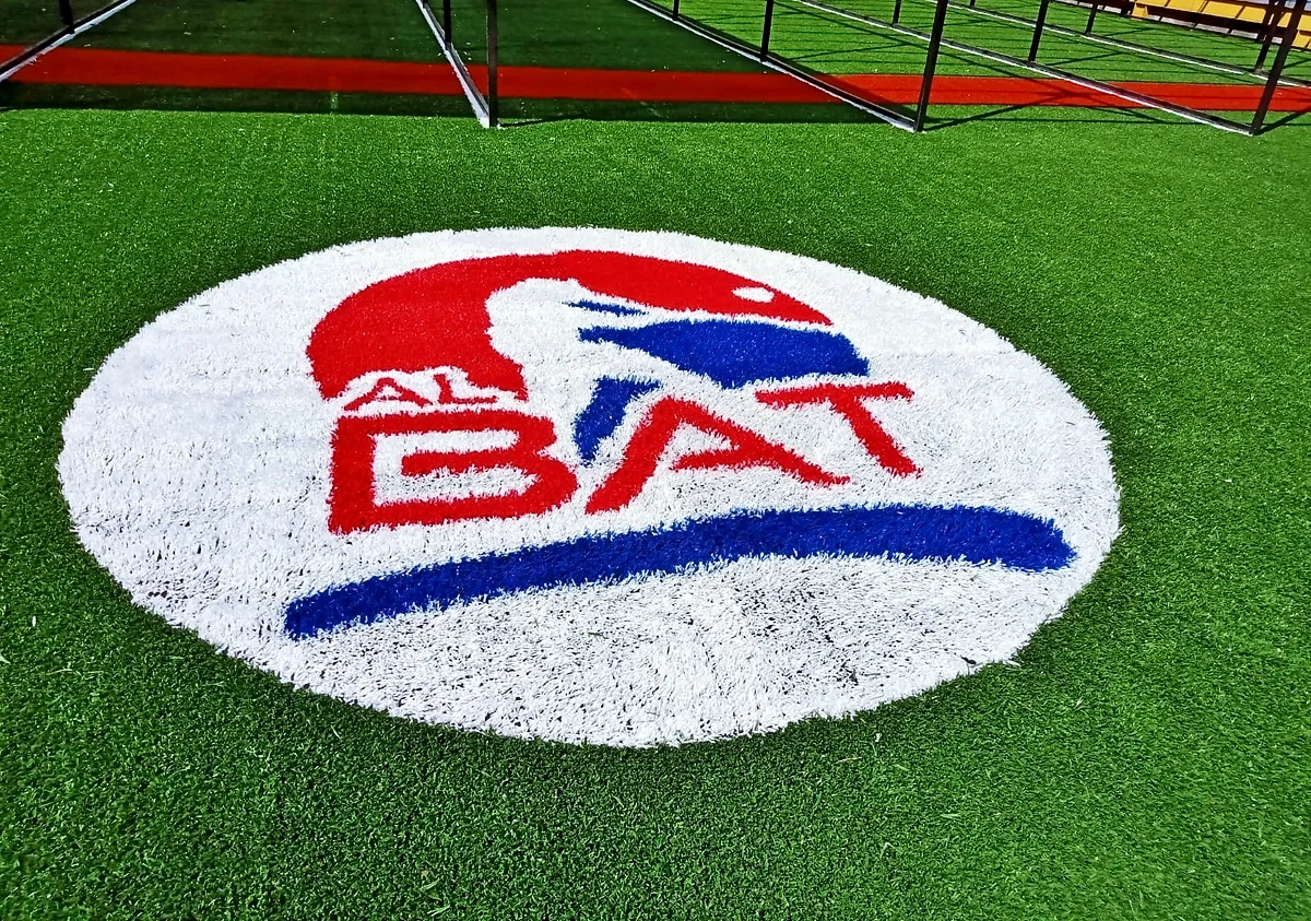 Detalle de cerca del logotipo AL BAT insertado en el pasto sintético, con la jaula de bateo al fondo