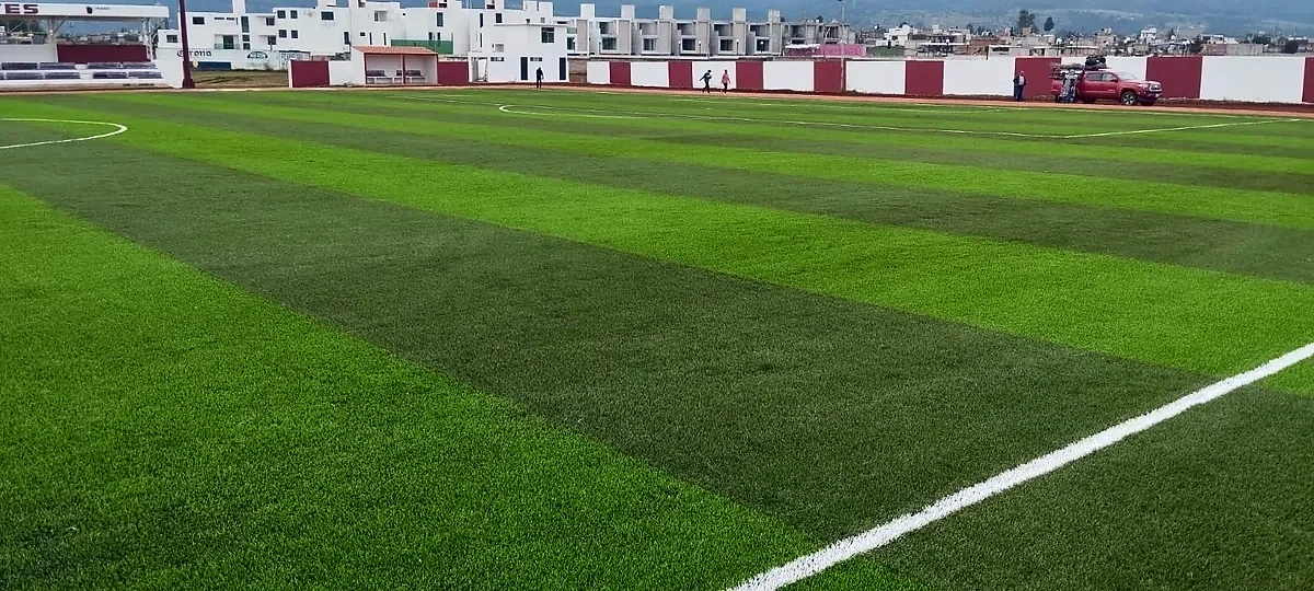 Franjas de dos tonos del pasto sintético de la cancha de soccer en Chignahuapan, Puebla, con el muro perimetral guinda y blanco